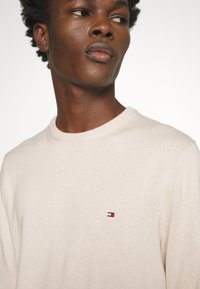 Maglione a maglia beige chiaro con scollatura rotonda, caratterizzato da un piccolo logo ricamato sul petto. Texture morbida, vestibilità normale.