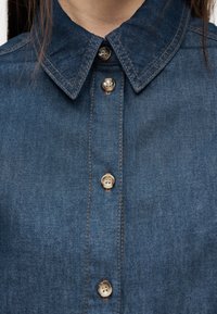 Suurelt võetud pilt inimesest, kes kannab sinist teksariidest särki, millel on terav krae ja neli nähtavat kinnitatud pruuni nööpi.