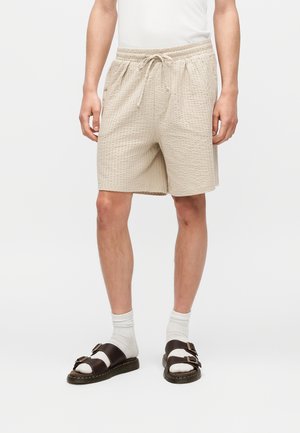 Férfi, aki bézs, texturált, húzózsinóros rövidnadrágot, fehér zoknit és sötétbarna, dupla pántos szandált visel, egyszínű háttér előtt áll.