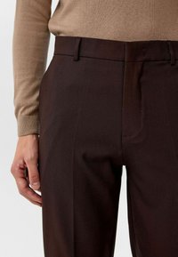 Donkerbruine op maat gemaakte broek met een gladde textuur en een platte voorkant. De tailleband heeft riemlussen, en de stof biedt een gestructureerde pasvorm.