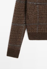 Brązowy sweter we wzory, wykonany z wełny, z fakturowaną powierzchnią, kieszenią i szarymi kratkami. Mankiet jest w ciemniejszym odcieniu brązu.