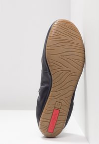 Semelle d'une chaussure en cuir noir avec une bande de roulement en caoutchouc beige, étiquette rouge « Kickers » et taille 37 indiquée en bas, inclinée sur un fond blanc.