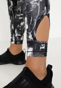 Černobílé legíny se vzorem a výřezem na lýtku, vyrobené z elastického materiálu; zkombinované s černými sportovními botami.