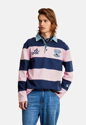 Jonge man met halflang haar die een marineblauw en roze gestreept rugbyshirt en blauwe spijkerbroek draagt, staand met één hand in zijn zak.