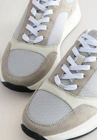 Hellgraue und beige Sneaker mit Mesh-Einsätzen, weißen Schnürsenkeln und einer schwarzen Sohle. Merkmale glatte und strukturierte Materialien in einem modernen Design.