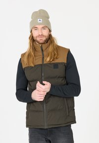 Padded vest with brown upper section and dark green lower section, featuring a zip closure and black logo patch. Black long-sleeve shirt underneath.