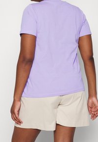 Person iført en lyse lilla kortærmet skjorte og beige shorts, vist fra ryggen mod en ensfarvet hvid baggrund.