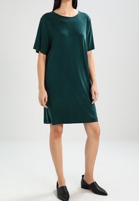 Robe verte à manches courtes, longueur au genou, avec un col rond, en tissu lisse. Associée à des chaussures plates noires à bouts pointus.