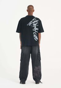 Camiseta negra de gran tamaño con un diseño en la parte de atrás que asemeja graffiti blanco. Combinada con pantalones cargo oscuros que presentan un bordado de dragón en la pierna.