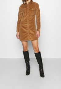 Korsettklänning i manchestertyg i beige, knälång, med två bröstfickor, knapplåst fram och långa ärmar. Matchad med svarta knähöga stövlar.