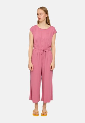 Junge Frau in einem lockeren rosa Jumpsuit mit einem Kordelzugbund und orangefarbenen Flip-Flops, die mit entspannten Armen dasteht und nach links schaut.