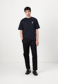 T-shirt en coton bleu marine à manches courtes et col rond, arborant un petit graphique sur la poitrine ; associé à un pantalon noir et des chaussures.