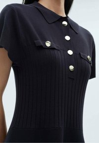 Robe en maille côtelée bleu marine avec col, manches courtes et poches avant ornées de boutons métalliques, mettant en valeur la texture et la coupe ajustée.