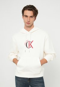 Hanorac alb din bumbac, cu buzunar frontal, glugă cu șnur și un logo Calvin Klein roșu și negru proeminent pe piept.