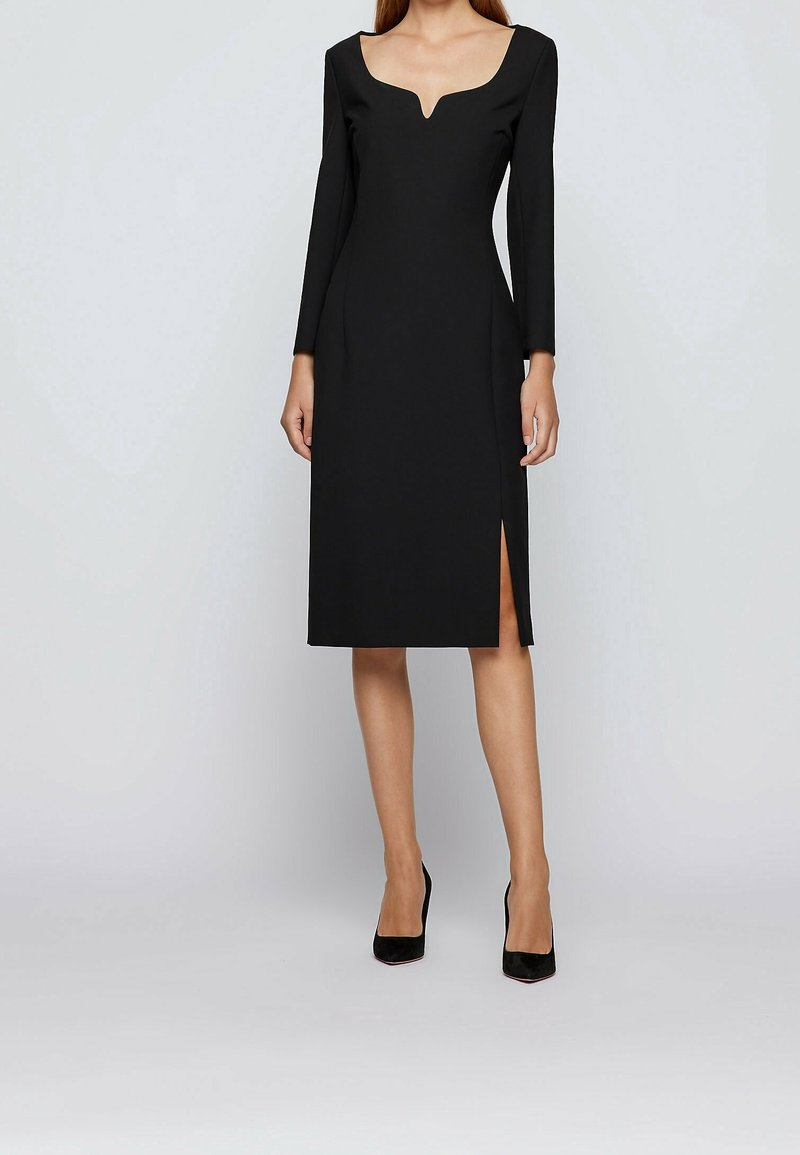 Femme portant une robe midi noire à manches longues avec un décolleté carré et une fente à l'avant, associée à des talons noirs à bout pointu, debout sur un fond neutre.