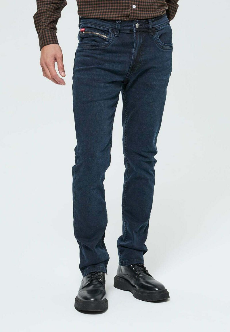 Homme portant un jean slim-fit bleu foncé, des chaussures noires à lacets et une chemise à carreaux marron, se tenant sur un fond blanc uni.