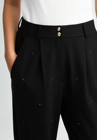 Pantaloni neri con un design sartoriale, caratterizzati da due bottoni dorati, tasche laterali e motivi ovali ricamati con accenti neri.