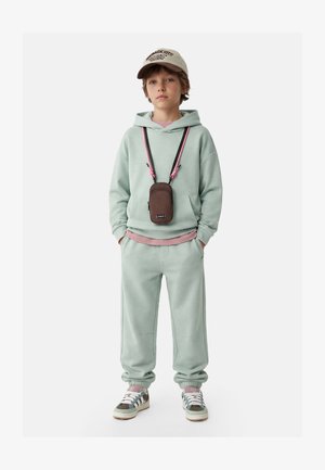 Kind draagt een lichtgroene hoodie en joggingbroek, beige pet, sneakers en een bruine crossbody tas met roze en zwarte bandjes.