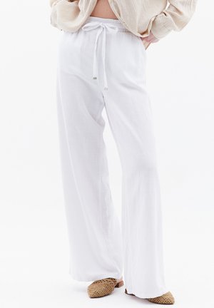Persona vistiendo pantalones blancos de pierna ancha con cintura ajustable con cordón y zapatos beige tejidos tipo slip-on sobre un fondo blanco.