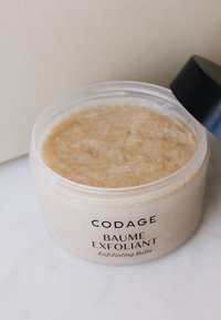 Peelingbalsam in einem klaren Glas mit schwarzem Deckel. Cremige beige Textur mit winzigen Peelingpartikeln. Etikett mit "CODAGE BAUME EXFOLIANT."