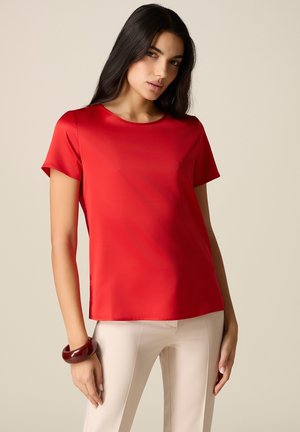 Donna con lunghi capelli scuri che indossa una camicetta rossa a maniche corte, pantaloni beige e un braccialetto marrone scuro sul polso sinistro, in piedi davanti a uno sfondo semplice.