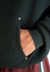 Felpa verde scuro con polsini a coste, caratterizzata da una tasca frontale fissata da bottoni a pressione argentati. La texture sembra morbida e accogliente.