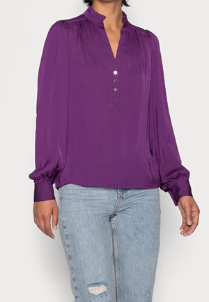 Kobieta w fioletowej bluzce z długim rękawem z falbaną na kołnierzu i trzema guzikami, w połączeniu z jasnoniebieskimi przetartymi jeansami.