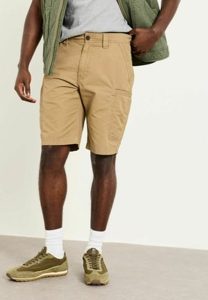 Mann trägt beige Cargo-Shorts, weiße Socken, olivgrüne Turnschuhe, graues Hemd und grüne gesteppte Weste, steht drinnen auf grauem Boden.