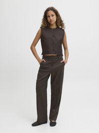 Colete sem mangas, em tom castanho com riscas finas, com botões à frente, combinado com calças largas a condizer. Modelo usa sapatilhas pretas texturizadas.