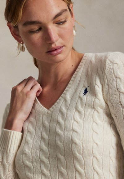 Femme portant un pull en maille torsadée crème à col en V avec un logo brodé bleu marine sur la poitrine, tenant le col et regardant vers le bas à sa gauche.