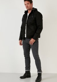 Svart jacka med huva, dragkedjestängning och texturerade axlar; tillsammans med mörkgrå jeans och svarta snörstövlar.