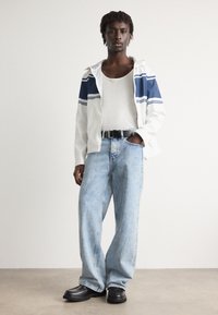 Hvid hættejakke med blå striber, grå tanktop, lyseblå baggy jeans og sorte sko. Glat stof og afslappet design.
