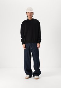 Schwarzes Sweatshirt, lockere dunkle Jeans in Blau und weiße Sneaker mit hellbrauner Sohle. Helle Kappe nach vorne getragen. Einfarbiger Hintergrund.