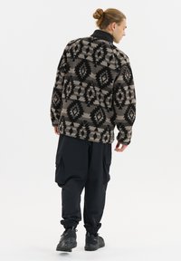 Fleece-Pullover in Beige mit schwarzen geometrischen Mustern, kombiniert mit schwarzen Cargo-Hosen und Stiefeln; verfügt über einen hohen Kragen und eine lockere Passform.