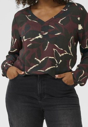 Femme portant un chemisier à col en V à motif floral foncé à manches longues, rentré dans un jean noir taille haute, les mains dans les poches.