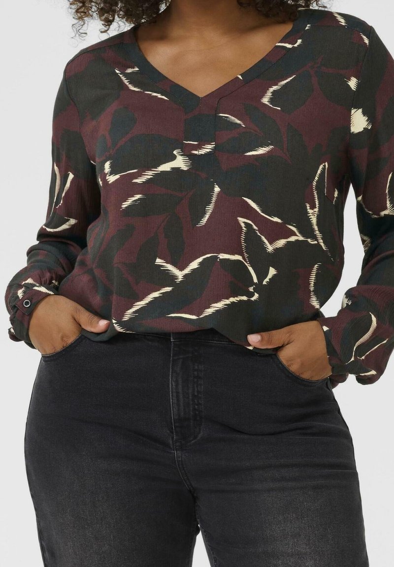 Femme portant un chemisier à col en V à motif floral foncé à manches longues, rentré dans un jean noir taille haute, les mains dans les poches.