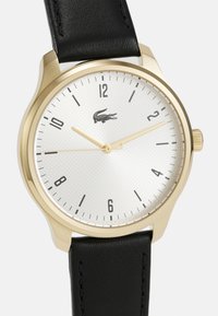 Reloj en tono dorado con una esfera texturizada blanca, marcadores de hora negros y una correa de cuero negro. Presenta manecillas doradas y un logo de Lacoste.