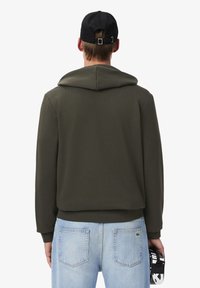 Sweat à capuche vert olive avec un cordon de serrage, texture douce et poignets côtelés. Associé à un jean bleu clair et une casquette noire.
