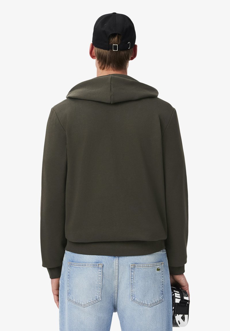 Olijfgroen hoodie met een trekkoord capuchon, zachte textuur en geribbelde manchetten. Gecombineerd met lichtblauwe jeans en een zwarte pet.