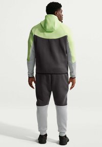 Homme de dos portant un sweat à capuche tricolore avec des panneaux vert lime, gris foncé et gris clair, assorti à un pantalon de jogging et des baskets noires.