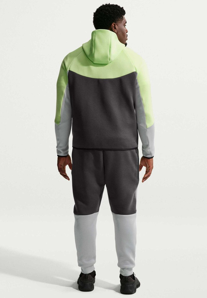 Mand set bagfra iført en tre-farvet hættetrøje med limegrønne, mørkegrå og lysegrå paneler, kombineret med matchende joggingbukser og sorte sneakers.