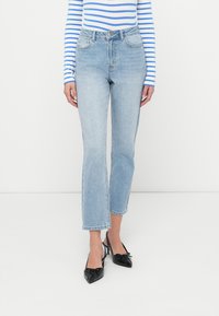 Φωτεινά γαλάζια τζιν με ψηλόμεση γραμμή και ίσια εφαρμογή, με ξέφτια στο τελείωμα· συνδυασμένα με μαύρο τακουνι με μυτερή μύτη και ριγωτό μακρυμάνικο τοπ.