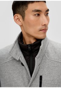 Grijze houndstooth jacket met een hoge kraag en een zwarte binnenlaag. Heeft een rits sluiting en een gestructureerde stof, met knopen op de kraag.