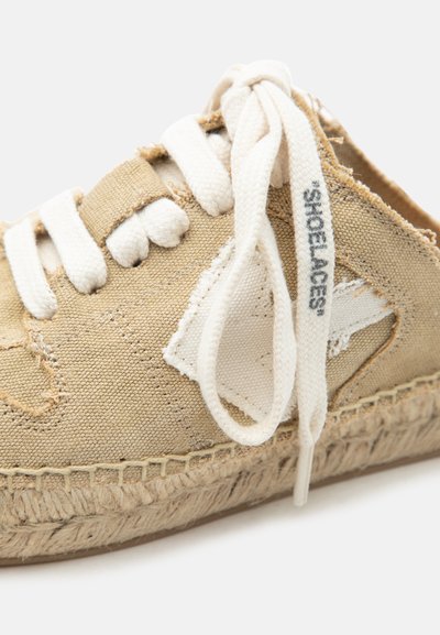 Κοντινό πλάνο μπεζ καμβά παπουτσιού με κορδόνια και πλεκτή σόλα, με την επιγραφή «SHOELACES» στα λευκά κορδόνια.