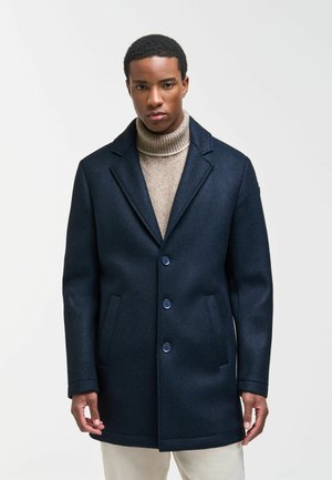 Junger Mann, der einen marineblauen knöpfbaren Mantel über einem beigen Rollkragenpullover mit hellen Hosen trägt, vor einem einfarbigen hellen Hintergrund stehend.