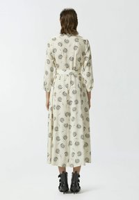 Lungo vestito beige con motivi neri a spirale, cintato in vita, con maniche lunghe e una linea fluida, abbinato a stivaletti neri texturizzati.