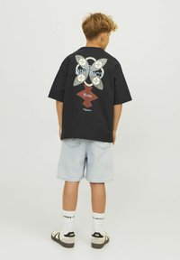 Svart bomullströja med en färgglad fjärilsdesign, denimshorts och svart-vita sandaler med strumpor, sedd bakifrån.