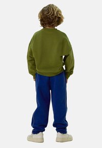 Sweatshirt en coton vert et joggers bleus avec des rayures blanches sur les côtés. Coupe décontractée, poignets côtelés et tissu texturé. Baskets blanches.