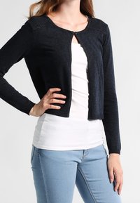 Cárdigan de punto azul marino oscuro con cuello redondo y cierre de botones, que presenta acentos texturizados en los hombros y mangas largas. Llevado sobre una blusa blanca.