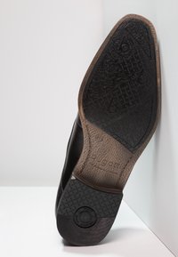 Suela de zapato de cuero negro con una superficie texturizada, que presenta el nombre de la marca "bugatti" y un área de agarre con un patrón en marrón y negro.